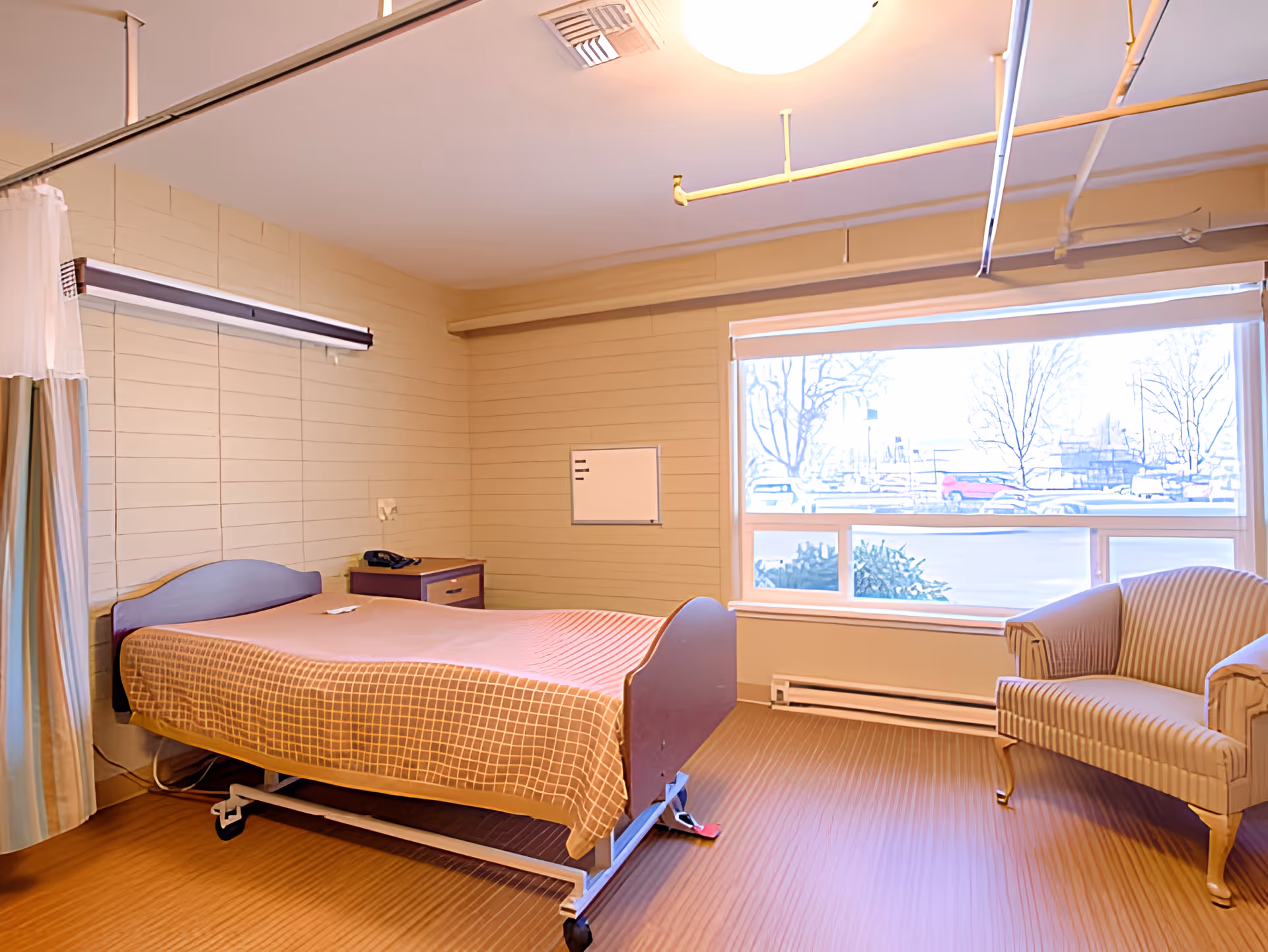 Well-lit care facility bedroom with a single adjustable bed, bedside table, armchair, and a large window.