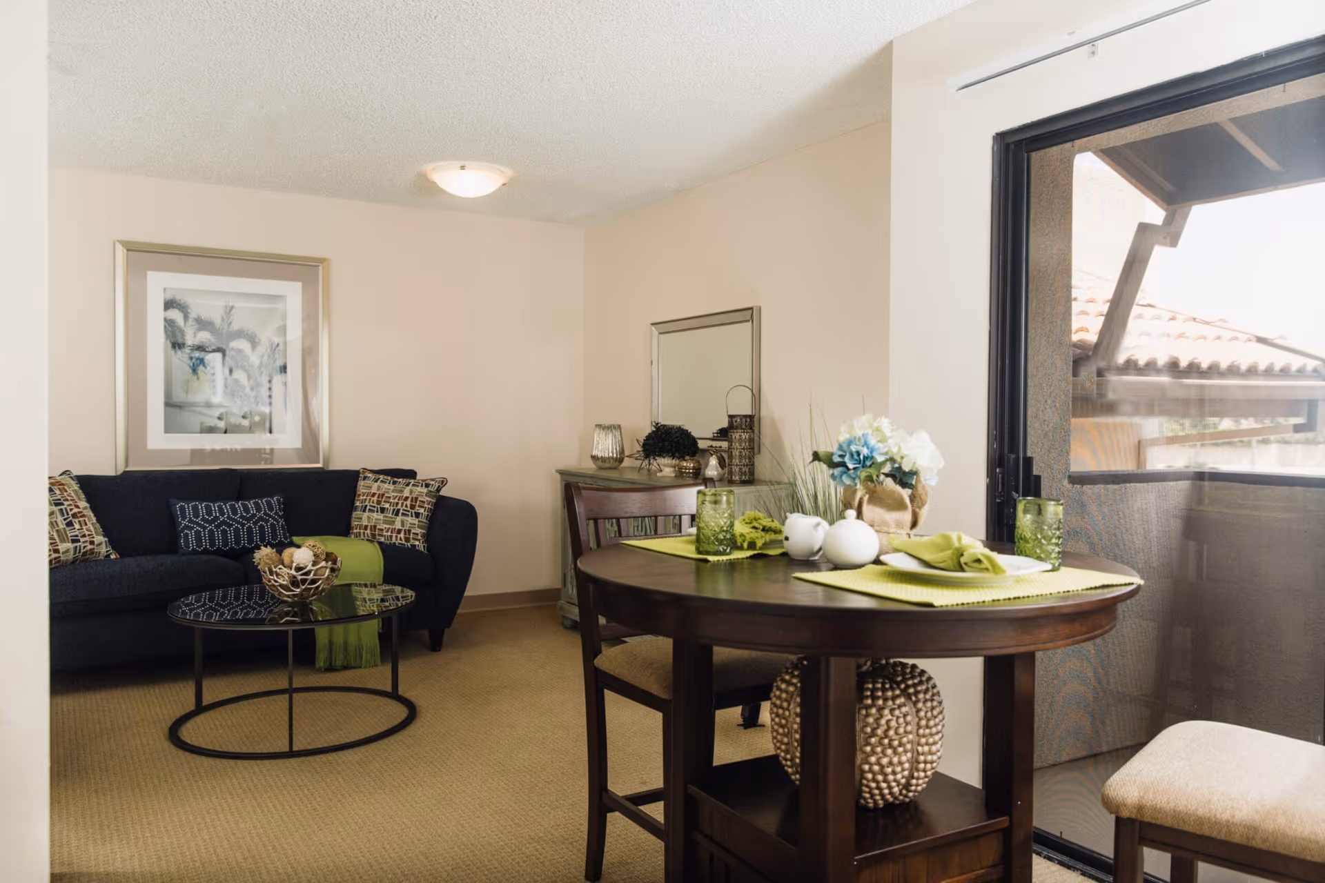 A cozy senior living suite interior showing a round dining table set with place settings by a sliding glass door and a sofa with a coffee table in the background.