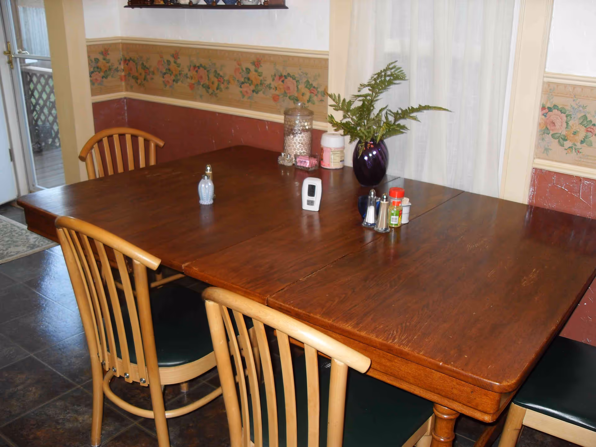 A wooden dining table with four wooden chairs around it. On the table, there are various items including a purple vase with green fern leaves, salt and pepper shakers, a small digital device, and some condiment containers. The room has floral wallpaper with a chair rail and a window with sheer white curtains in the background.