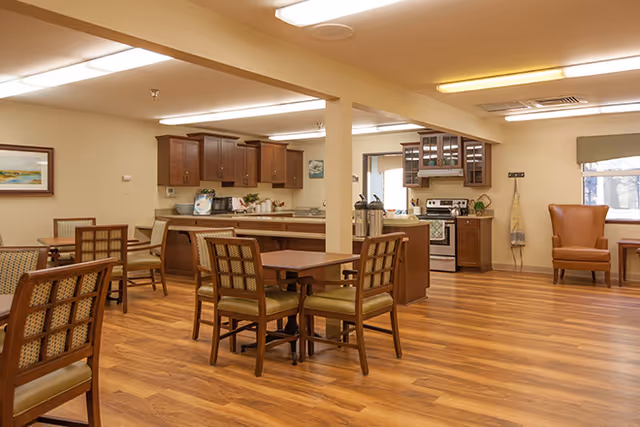 A bright communal dining room with multiple wooden tables and chairs, wood-look floors, and an open kitchen with cabinets and appliances.