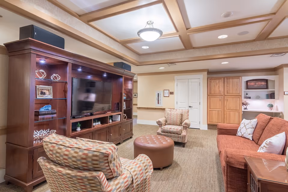 Comfortable communal living room with a sofa, patterned armchairs, round ottoman, and a large wooden entertainment center with a TV.