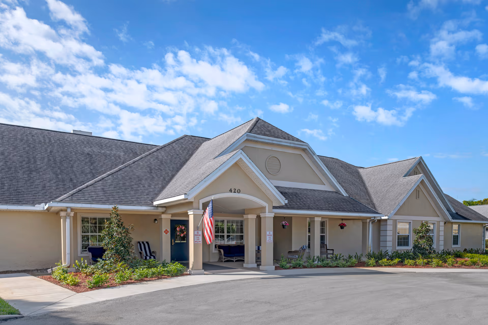 Front exterior of a single-story memory care building with a covered entryway, seating, landscaping, and an American flag.