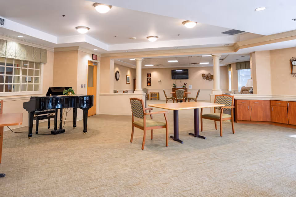 A spacious common area in a senior living facility featuring a black grand piano on the left, a wooden table with two chairs in the center, and another seating area with a table and chairs in the background. The room has beige walls, carpeted floors, recessed lighting, and large windows with valances letting in natural light.