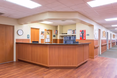 Reception desk area inside a healthcare facility with wood paneling and flooring, a clock on the wall, doors leading to other rooms, and a hallway extending to the right.
