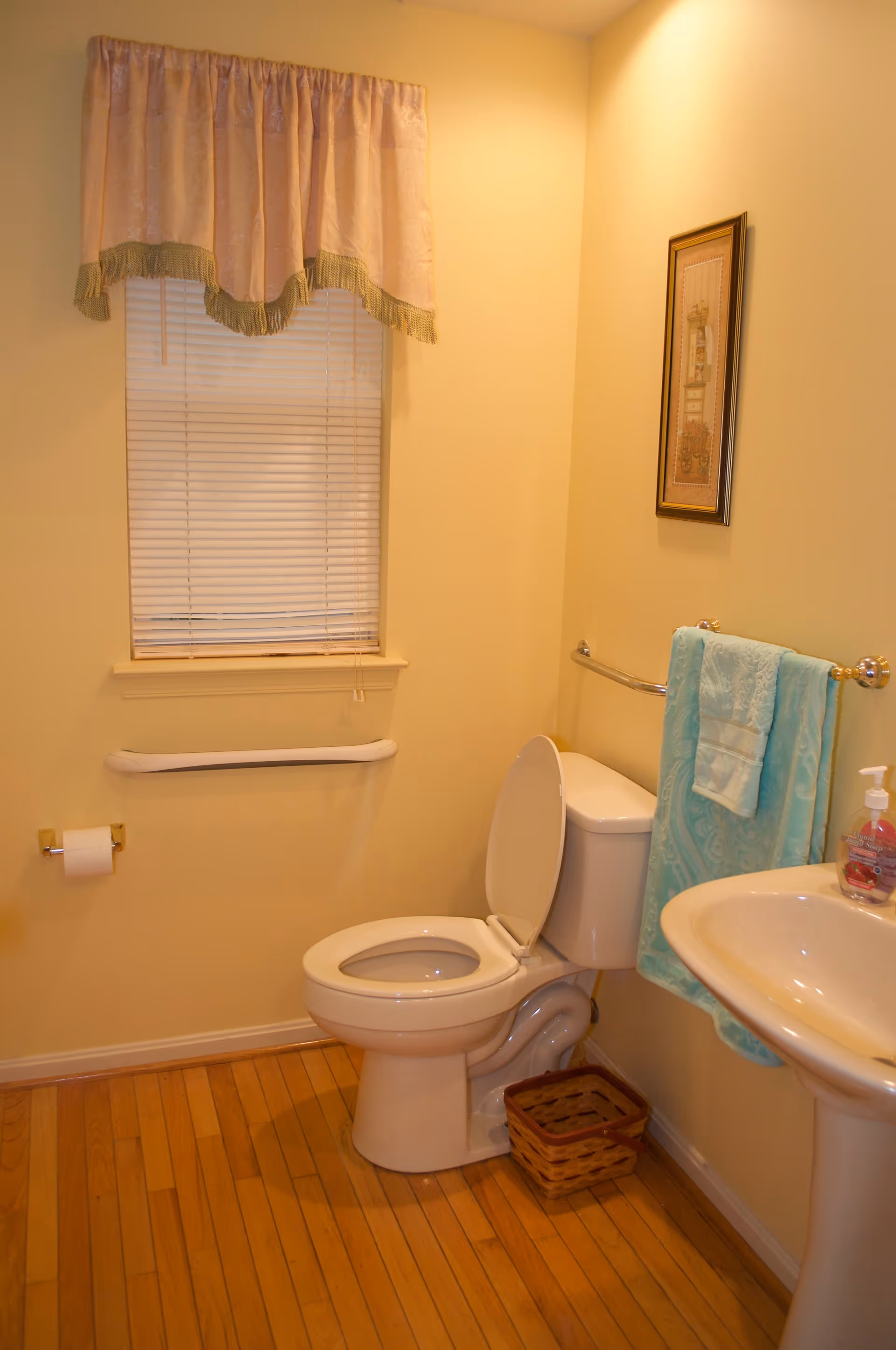 A small bathroom with a white toilet with the lid open, a white pedestal sink with a bottle of liquid hand soap, a towel rack with a turquoise towel, a window with closed blinds and a pink valance curtain, a framed picture on the wall, a toilet paper holder with a roll of toilet paper, a grab bar on the wall, and a small wicker basket on the wooden floor.