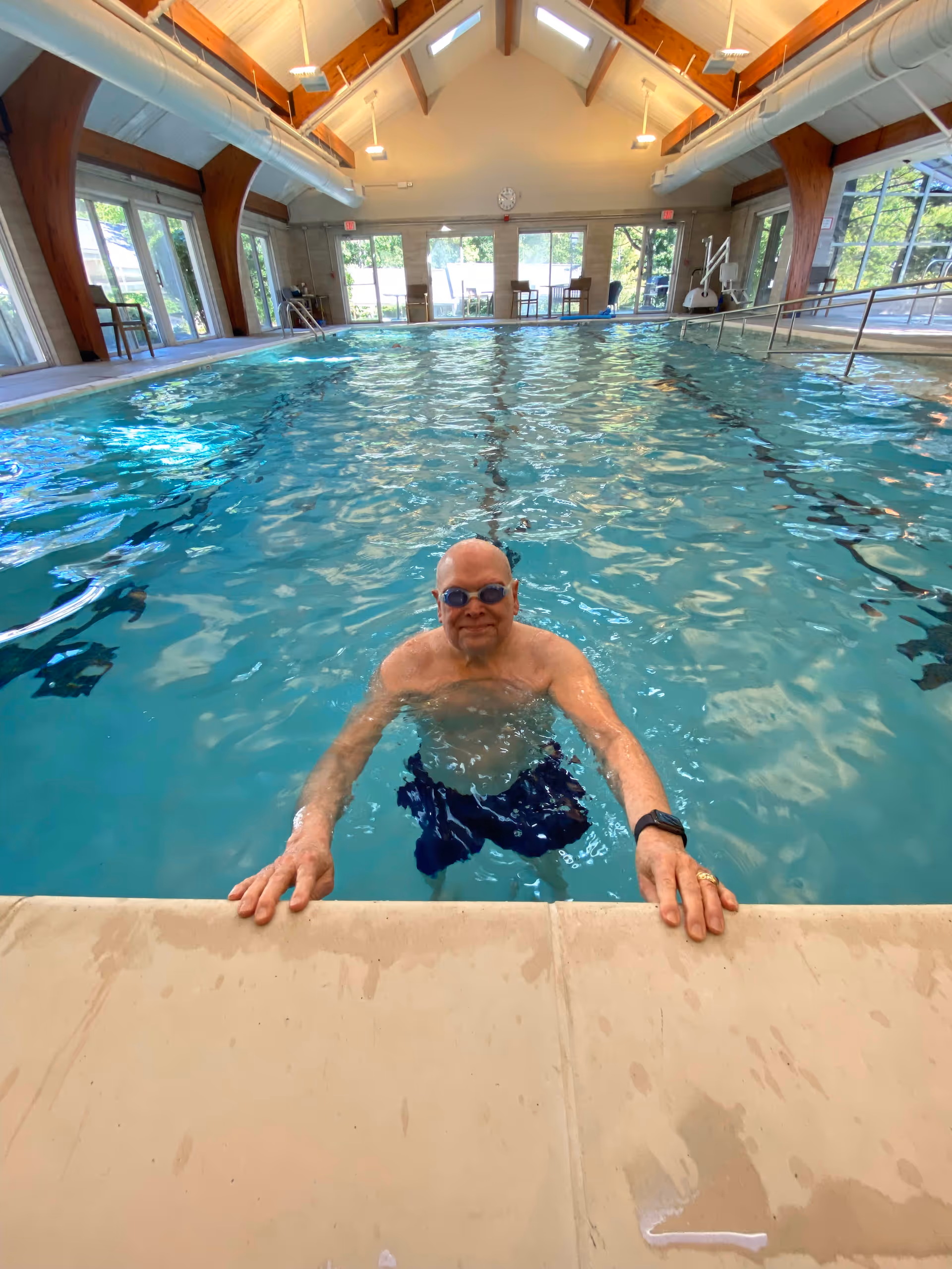 An elderly man wearing swimming goggles and swim trunks is standing in an indoor swimming pool, holding onto the edge. The pool area has large windows letting in natural light, wooden beams supporting the ceiling, and chairs placed along the sides.
