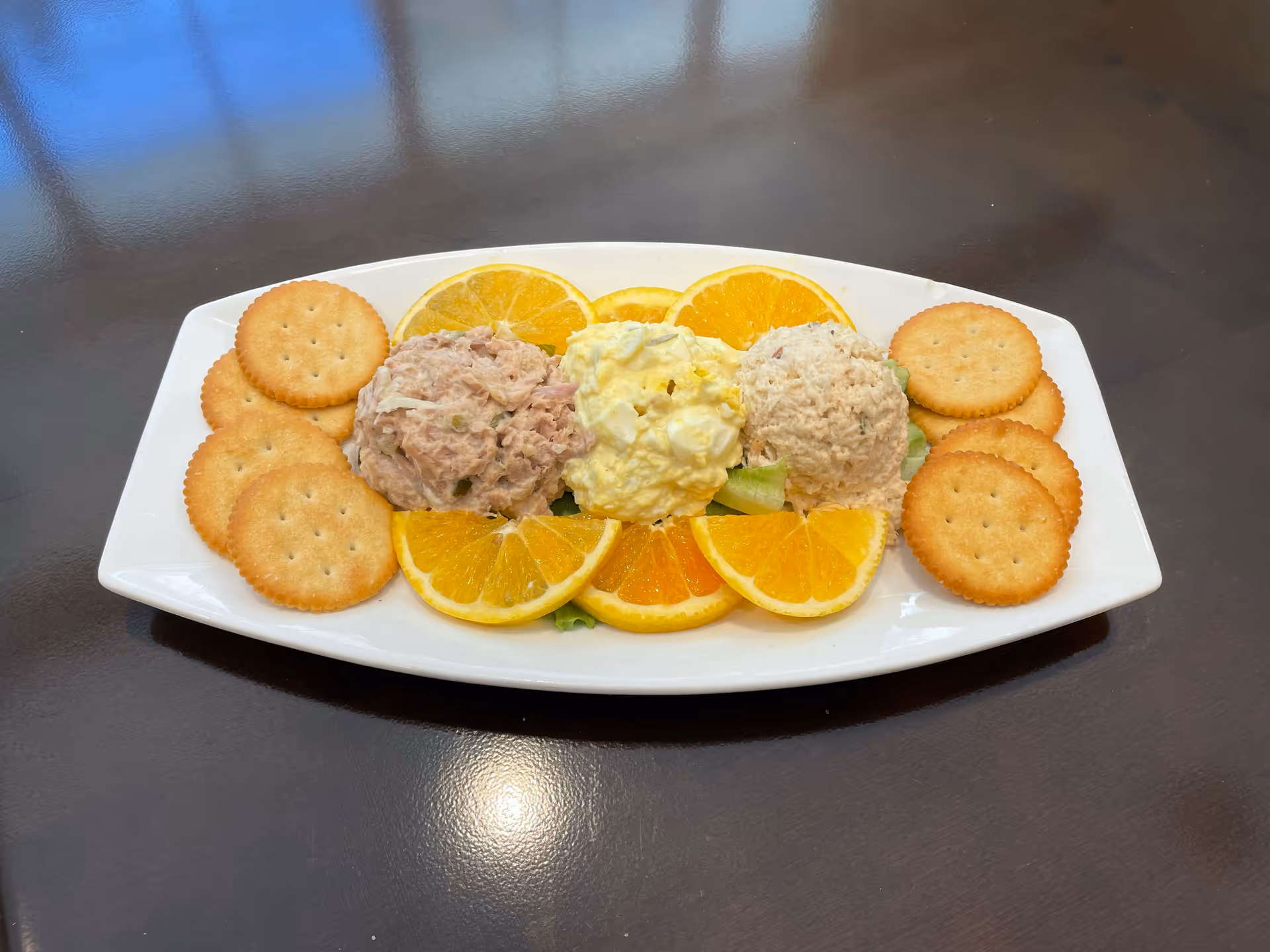A white platter with three scoops of salad (tuna, egg, and chicken) garnished with orange slices and round crackers on a dark table.