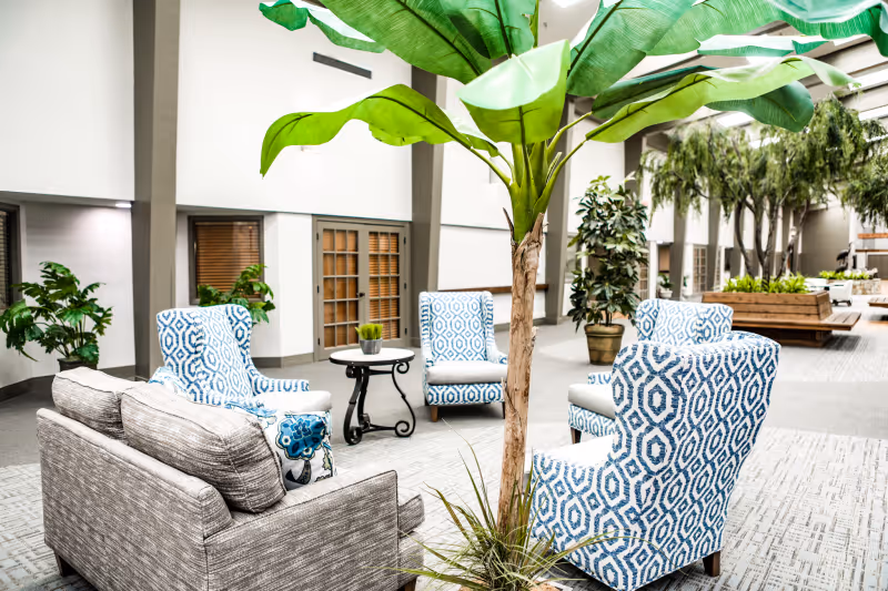Indoor atrium-style lounge with patterned blue armchairs, a gray sofa, side tables, and large potted plants.