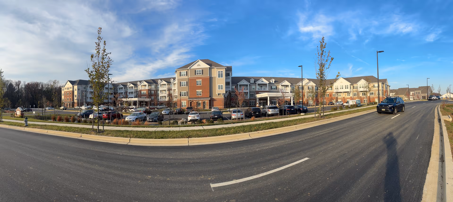Wide exterior view of a multi-story senior living facility building with a parking lot and a road in front under a blue sky.