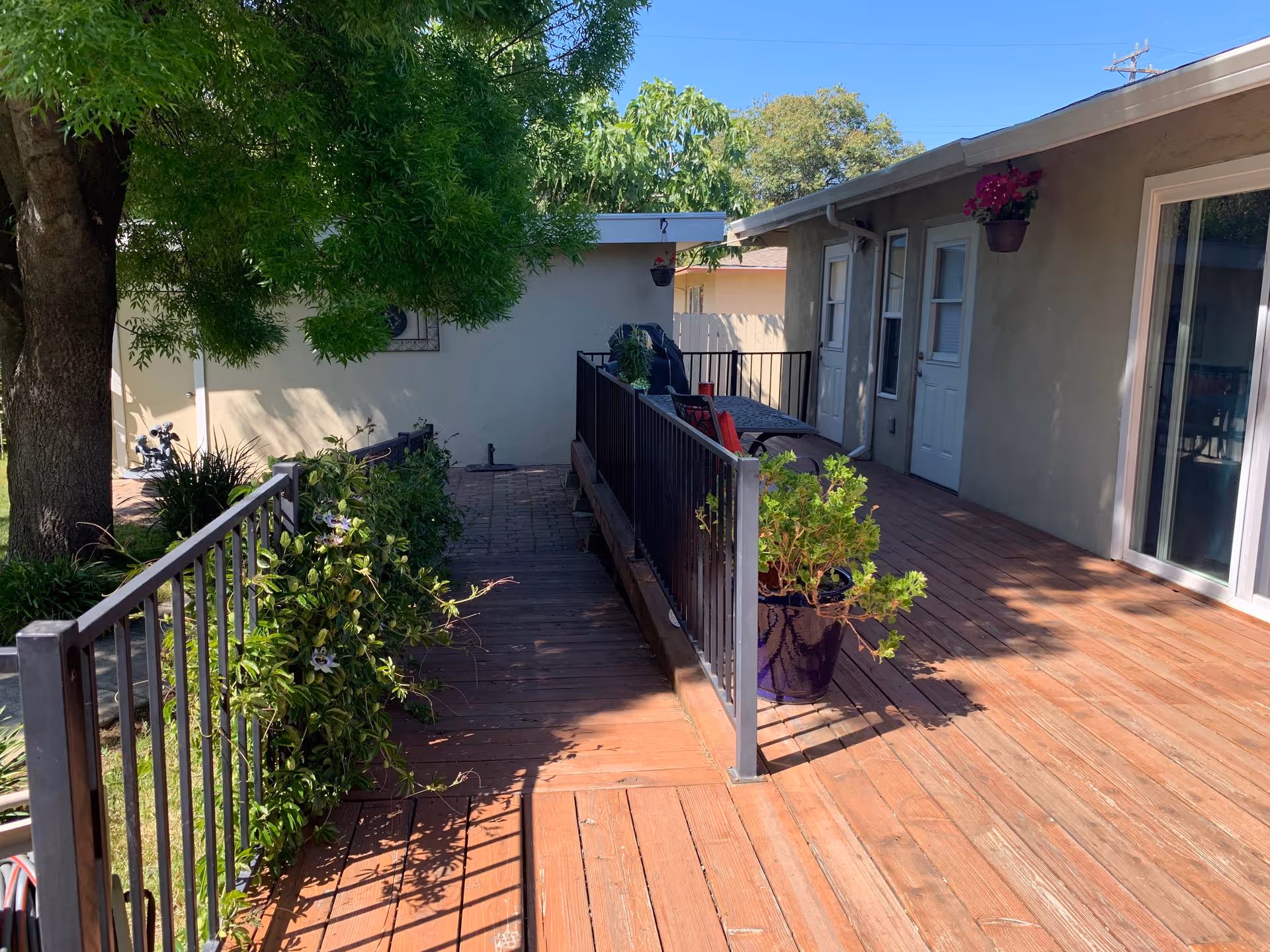 Wooden outdoor deck with a ramp and metal railing, potted plants, and doors leading into a single-story building under a clear blue sky.