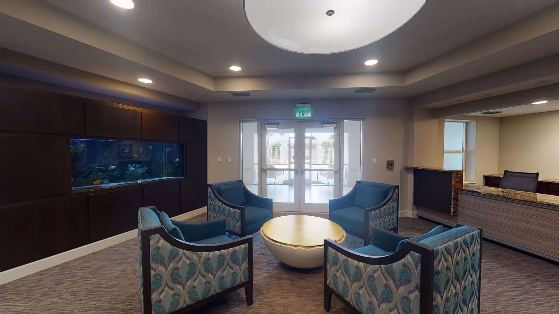 A cozy seating area in a memory care community with four patterned armchairs arranged around a round coffee table. There is a large built-in aquarium on the left wall and a reception desk on the right. Double glass doors lead to an outdoor area, and the ceiling features recessed lighting and a circular light fixture.