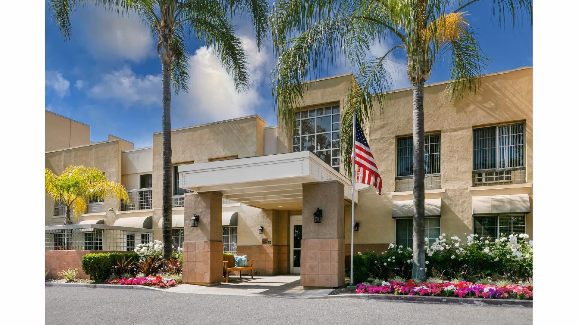 Exterior view of a senior living facility named Park Plaza with a covered entrance, palm trees, an American flag, and colorful flower beds under a partly cloudy sky.