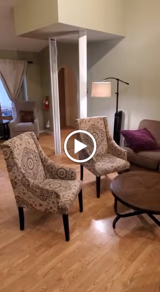 A cozy living room area with two patterned armchairs facing a round wooden coffee table. In the background, there is a beige sofa with a purple throw pillow, a floor lamp, and a small table with a chair near a window with curtains. The room has light-colored wooden flooring and light green walls.
