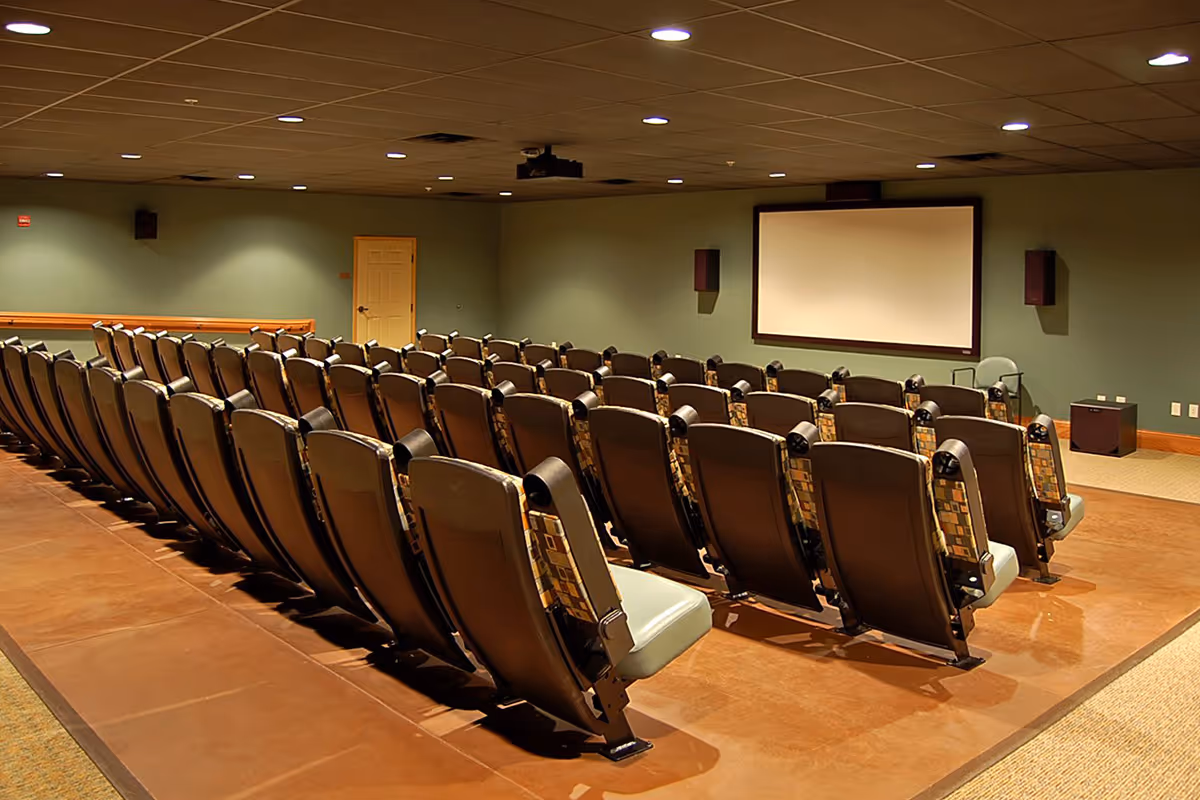 A small auditorium or theater room with several rows of cushioned seats facing a large blank projection screen on the wall. The room has a brown floor, green walls, and a ceiling with recessed lighting. There is a door at the back and speakers mounted on the walls.