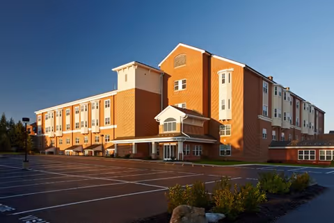 Exterior view of a large multi-story senior living facility building with a spacious empty parking lot in the foreground and some landscaping with bushes and rocks near the entrance, under a clear blue sky.