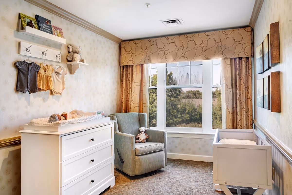 Nursery room with a white changing dresser, gray upholstered armchair, crib, wall-mounted shelves holding baby clothes and toys, and a large window with patterned curtains.