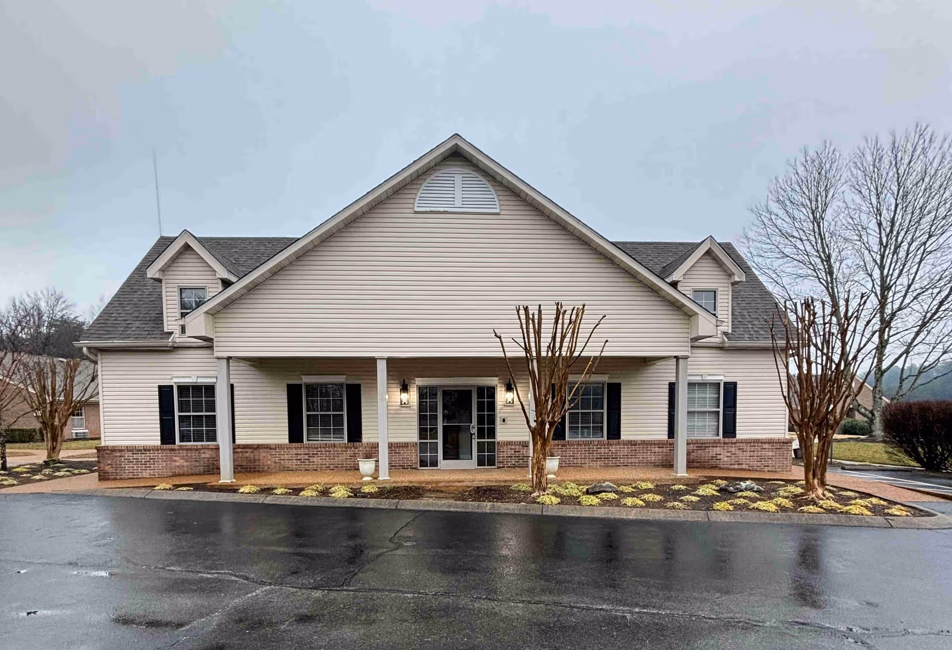 Front exterior of a beige senior living building with a covered entrance, columns, trimmed trees, and a wet driveway.