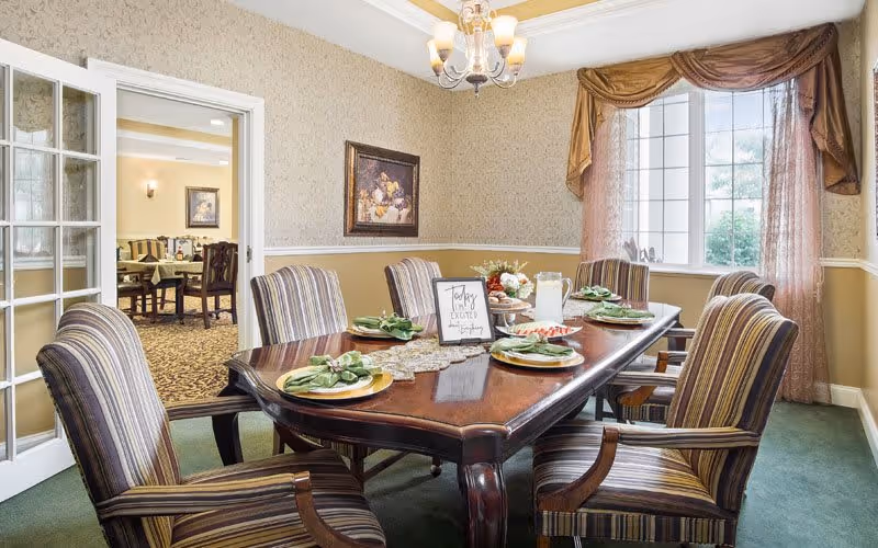 A formal dining room with a polished wooden table set for six people with plates, napkins, and a centerpiece. The room has striped upholstered chairs, a chandelier overhead, beige wallpaper, and a large window with sheer curtains and a valance. Through a glass-paneled door, another dining area with tables and chairs is visible.