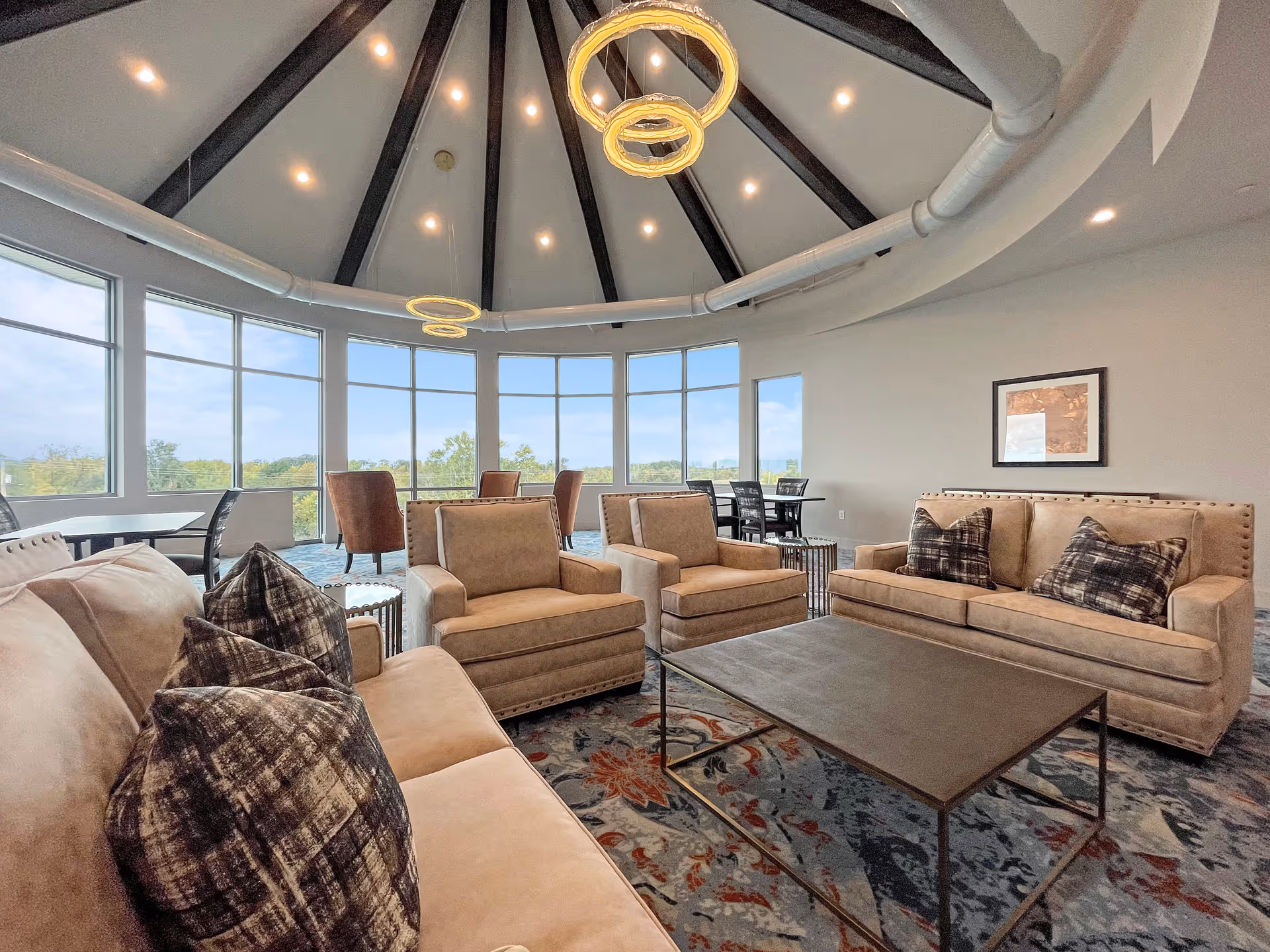 Circular lounge with beige sofas and armchairs around a coffee table, large windows overlooking trees, and a domed ceiling with ring chandeliers.