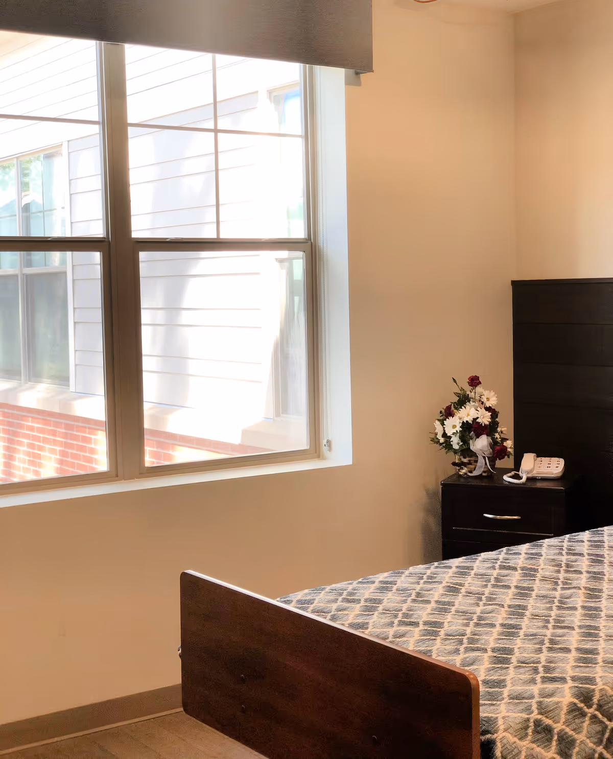 A bright bedroom corner with a large window letting in natural light. The room features a wooden bed with a patterned bedspread, a dark wooden nightstand with a floral arrangement and a white telephone on top.