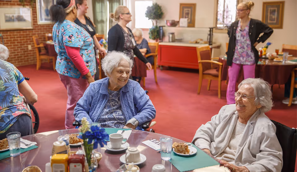 Two elderly women sitting at a round dining table with plates of food and drinks, smiling and engaging with each other. In the background, several caregivers and other elderly residents are standing and interacting in a warmly lit room with red carpet and wooden chairs.
