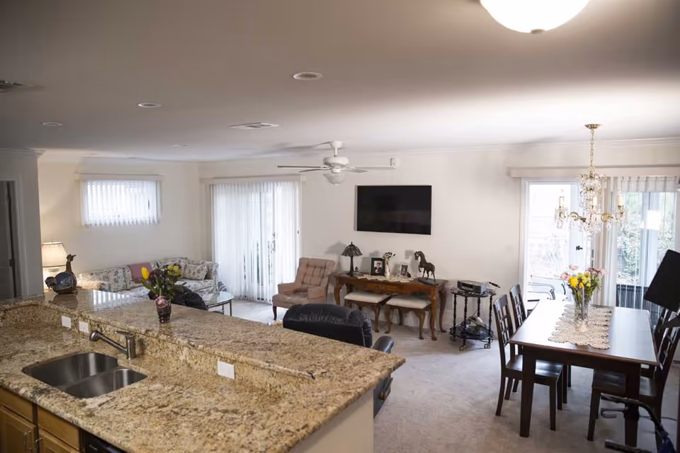 A bright and spacious living and dining area in a retirement community. The foreground features a granite countertop with a sink and faucet. Beyond the counter, there is a living room with a floral-patterned sofa, a beige armchair, and a flat-screen TV mounted on the wall above a wooden console table with decorative items. To the right, a dining table with six chairs is set under a chandelier, with a vase of flowers as a centerpiece. Large windows and sliding glass doors with vertical blinds allow natural light to fill the room.