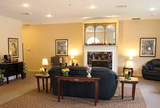 A cozy living room area in a senior living facility featuring a dark green leather sofa and armchair, a fireplace with a marble surround and decorative mirrors above it, two side tables with lamps and floral arrangements, framed artwork on the walls, and soft overhead lighting.