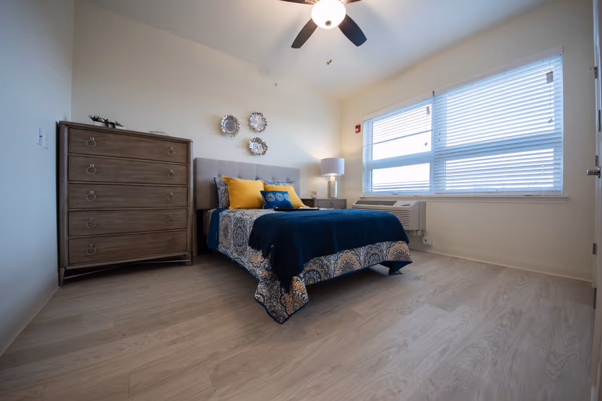A bright bedroom with a bed dressed in blue and patterned bedding, accented with yellow pillows. There is a wooden dresser to the left of the bed, a nightstand with a lamp to the right, and a large window with blinds letting in natural light. The room has light wood flooring and a ceiling fan with a light fixture.