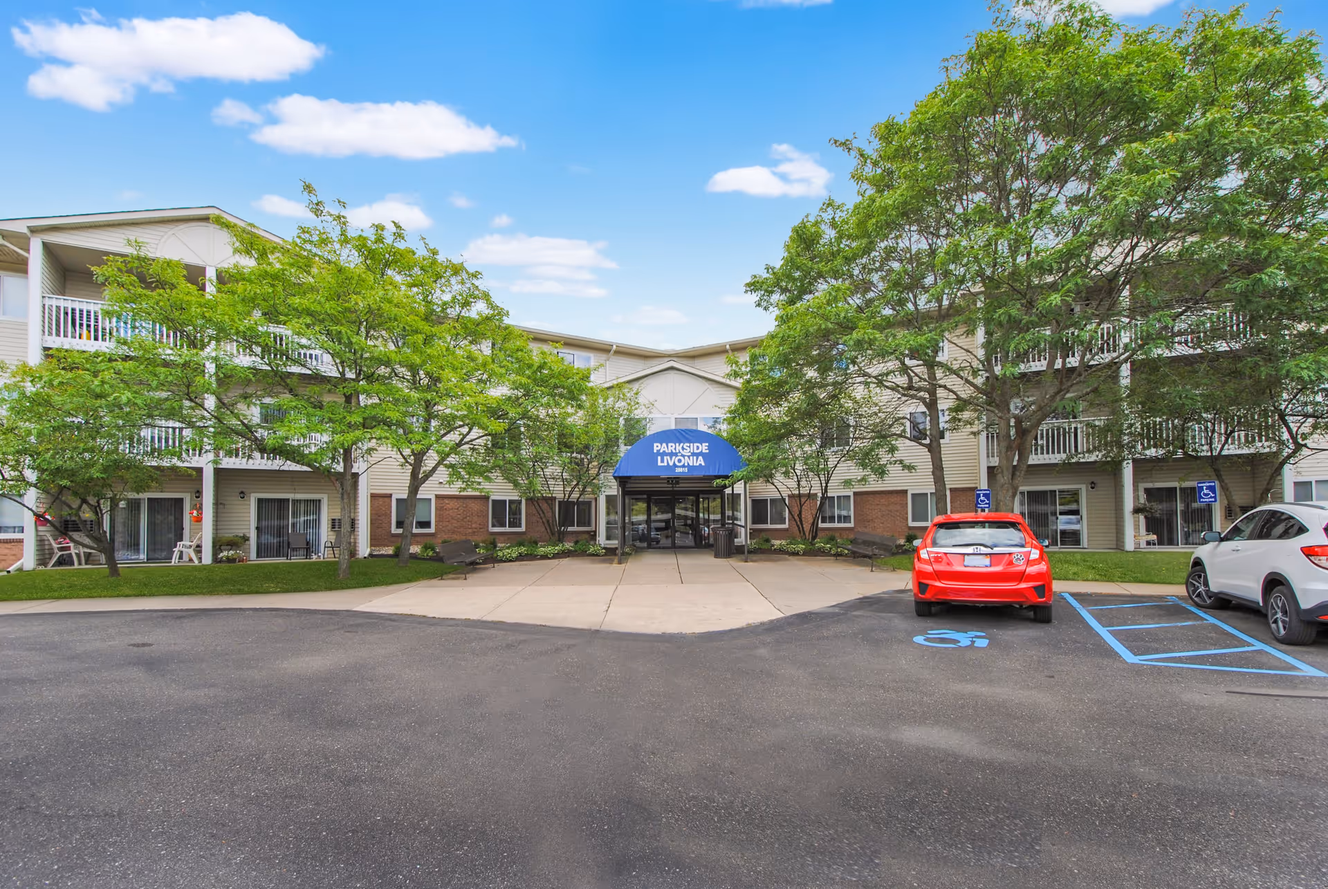 Front exterior view of Parkside of Livonia, a multi-story senior living facility with a blue awning over the main entrance. The building is surrounded by trees and has a parking lot with handicap parking spaces and cars parked.