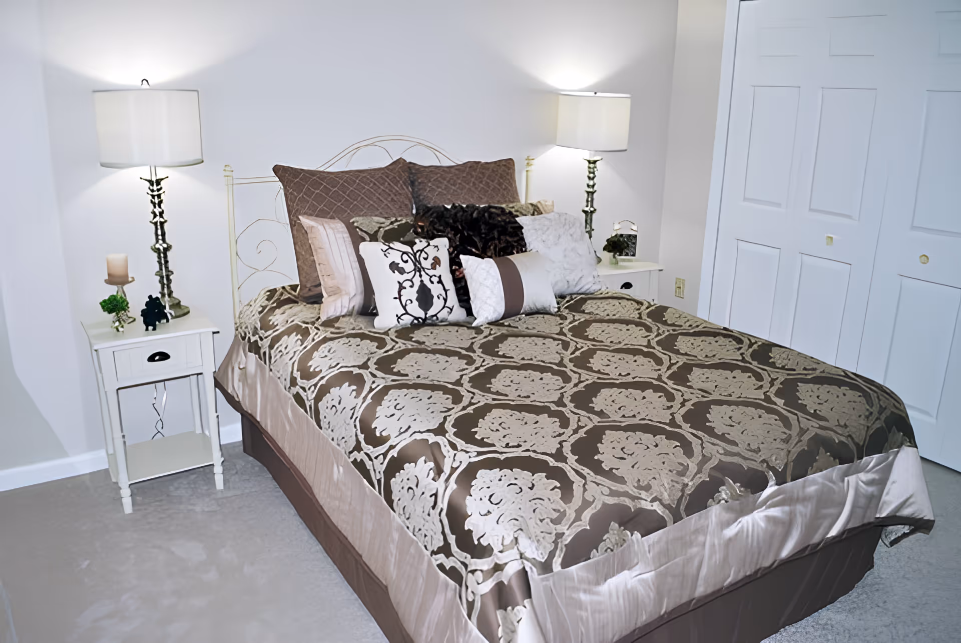 A neatly made bed with a decorative brown and beige patterned bedspread and multiple pillows in various colors and textures. The bed has a white metal headboard and is flanked by two white nightstands, each with a lamp. The room has light-colored walls and carpet, and there is a white closet door visible on the right side.