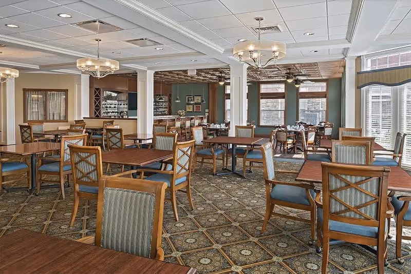 A spacious dining room with multiple wooden tables and chairs arranged neatly. The room features patterned carpet flooring, large windows with blinds allowing natural light, and elegant ceiling light fixtures. There is a bar area with shelves and glassware in the background.