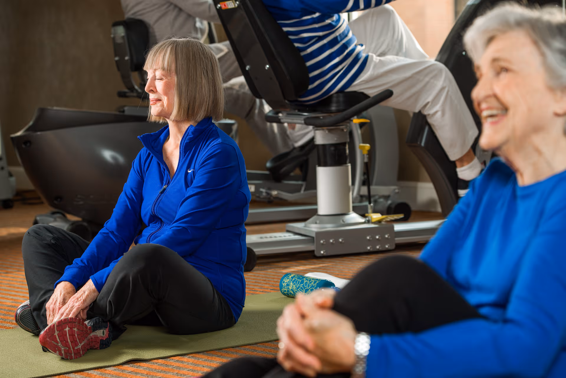 Two elderly women in blue tops sitting on exercise mats indoors, stretching and smiling, with exercise equipment in the background.