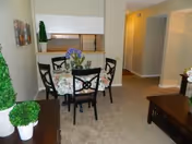 Interior view of a senior living apartment showing a small dining area with a round table covered by a floral tablecloth and four black chairs. Behind the dining area is a pass-through window to the kitchen. The room has beige carpet and walls, with a hallway leading to other rooms. There are decorative plants and artwork on the walls.