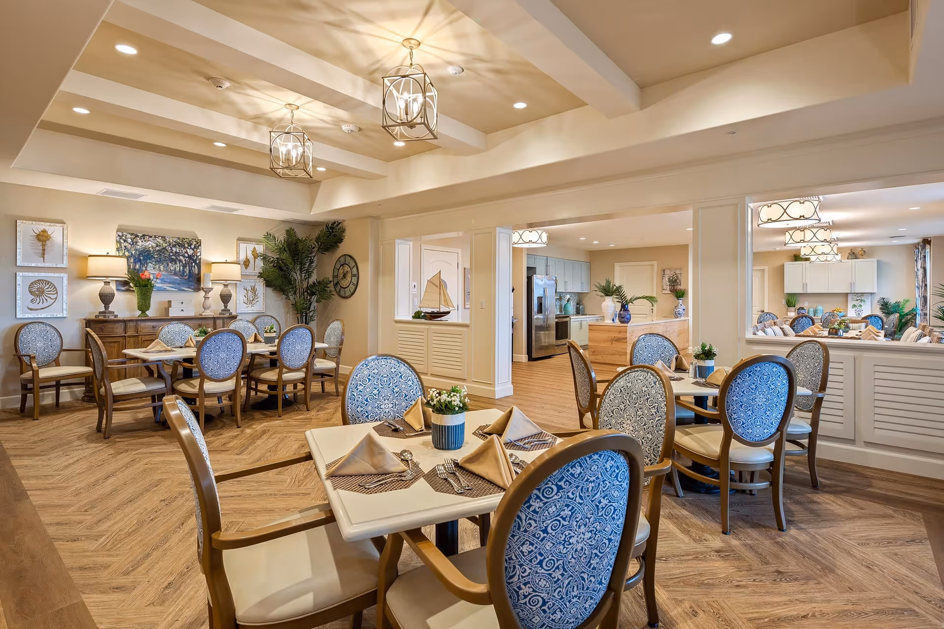 A bright and elegant dining room in a senior living facility with several tables set with beige napkins, silverware, and small flower arrangements. The chairs have wooden frames with blue patterned upholstery. The room features wooden flooring, decorative wall art, plants, and soft lighting from ceiling fixtures and table lamps. In the background, there is a kitchen area with stainless steel appliances and additional seating.