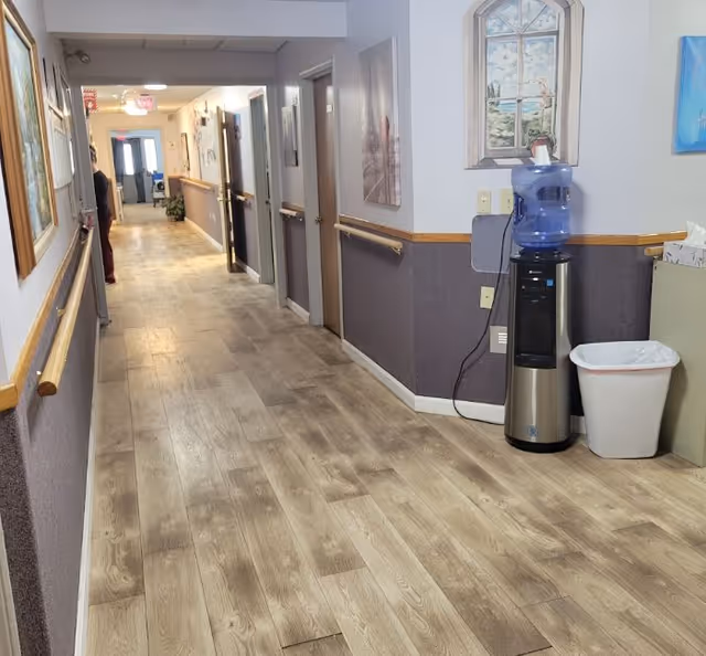 Well-lit interior hallway with wood-look flooring, handrails, several doors, wall art, and a water cooler with a trash bin.