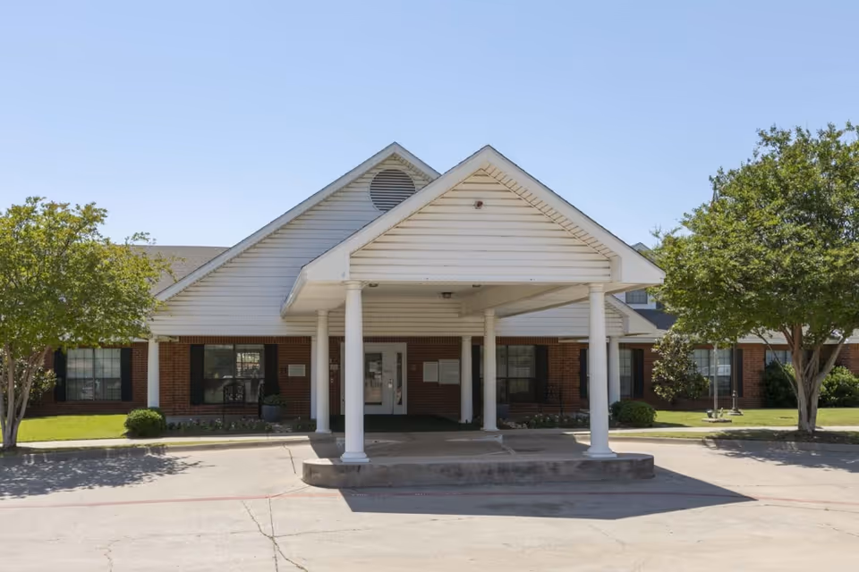 Front entrance of a single-story brick building with a white covered porte-cochère supported by columns and flanked by trees and windows.