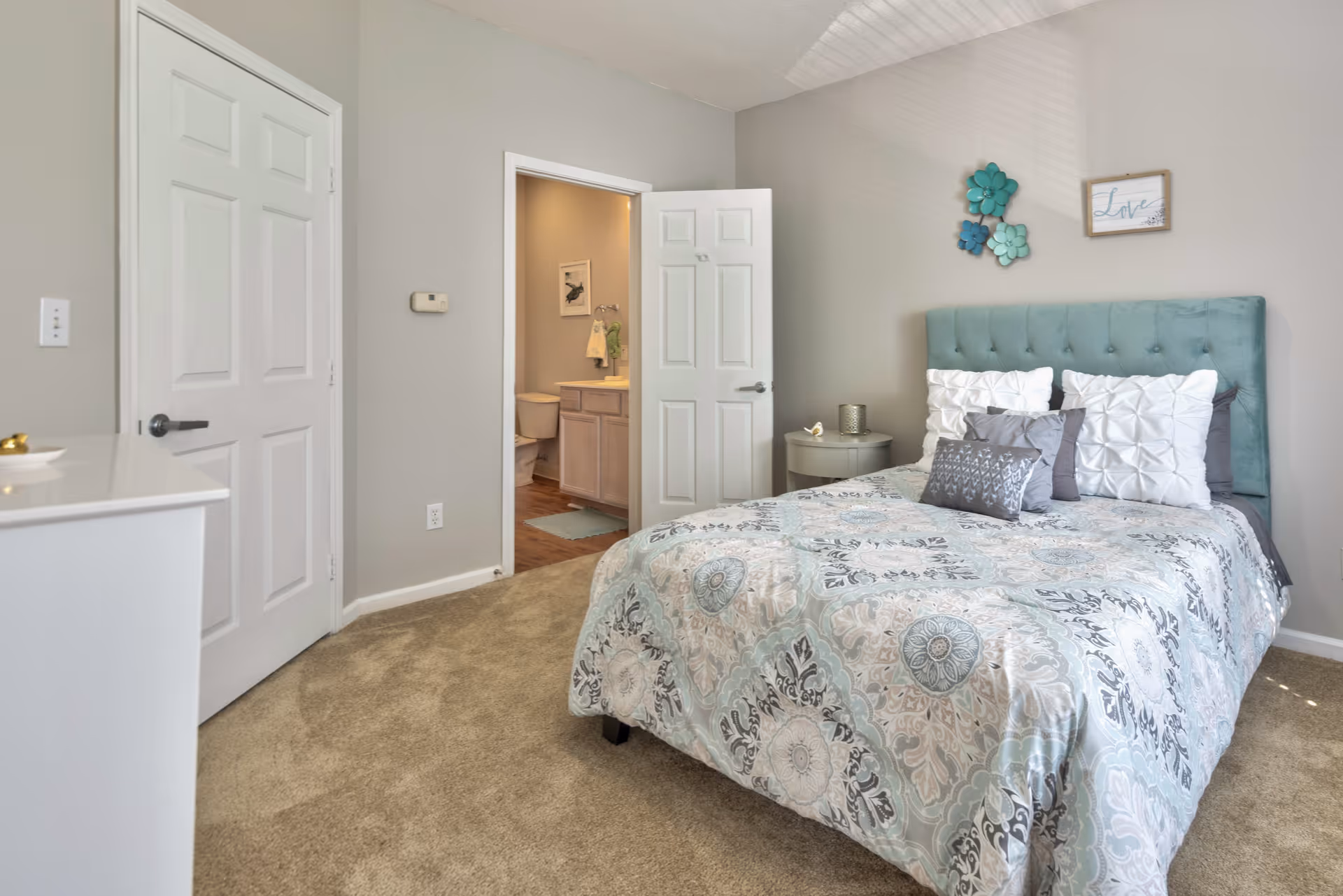 A bedroom with a teal upholstered headboard, a bed with patterned bedding and multiple pillows, a small round bedside table with a candle, and a dresser. The room has beige carpet and light gray walls. An open door reveals a bathroom with a toilet, sink, and a towel hanging on the wall.