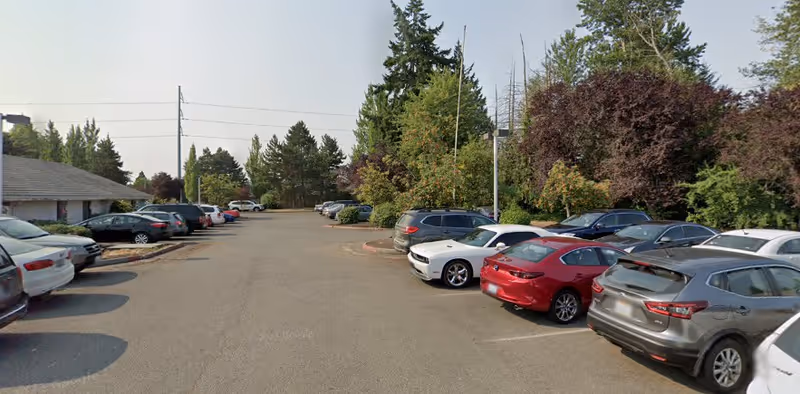 Parking lot with several parked cars on both sides, surrounded by trees and greenery under a clear sky.
