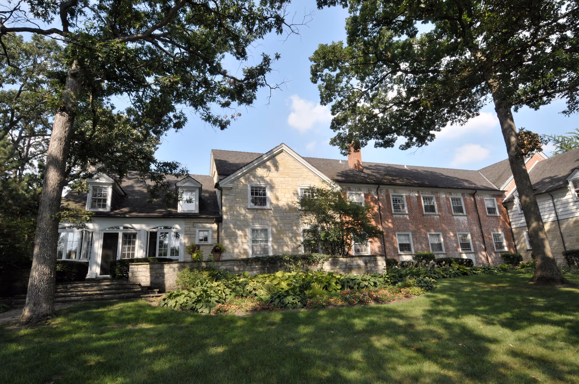 Exterior view of a large, multi-section building with stone and brick facades, surrounded by green grass, trees, and landscaped bushes under a blue sky with some clouds.