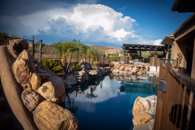 Outdoor swimming pool area surrounded by large rocks and landscaping with palm trees, a pergola with seating in the background, and a clear blue sky with clouds overhead.