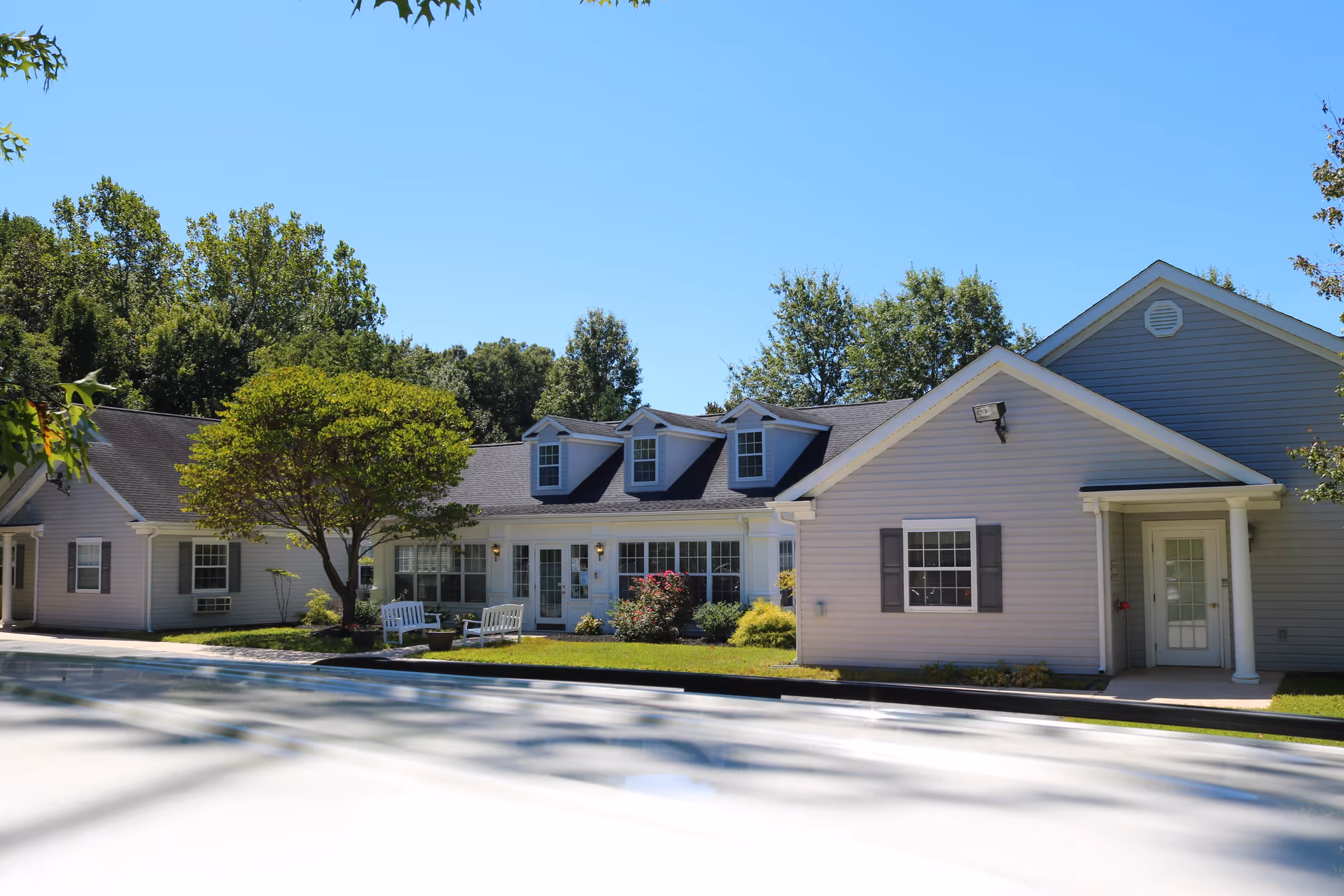 Exterior view of a single-story assisted living facility building with light gray siding, white trim, and multiple windows. There is a small tree and some bushes in front of the building, along with white outdoor benches on a grassy area. The sky is clear and blue.