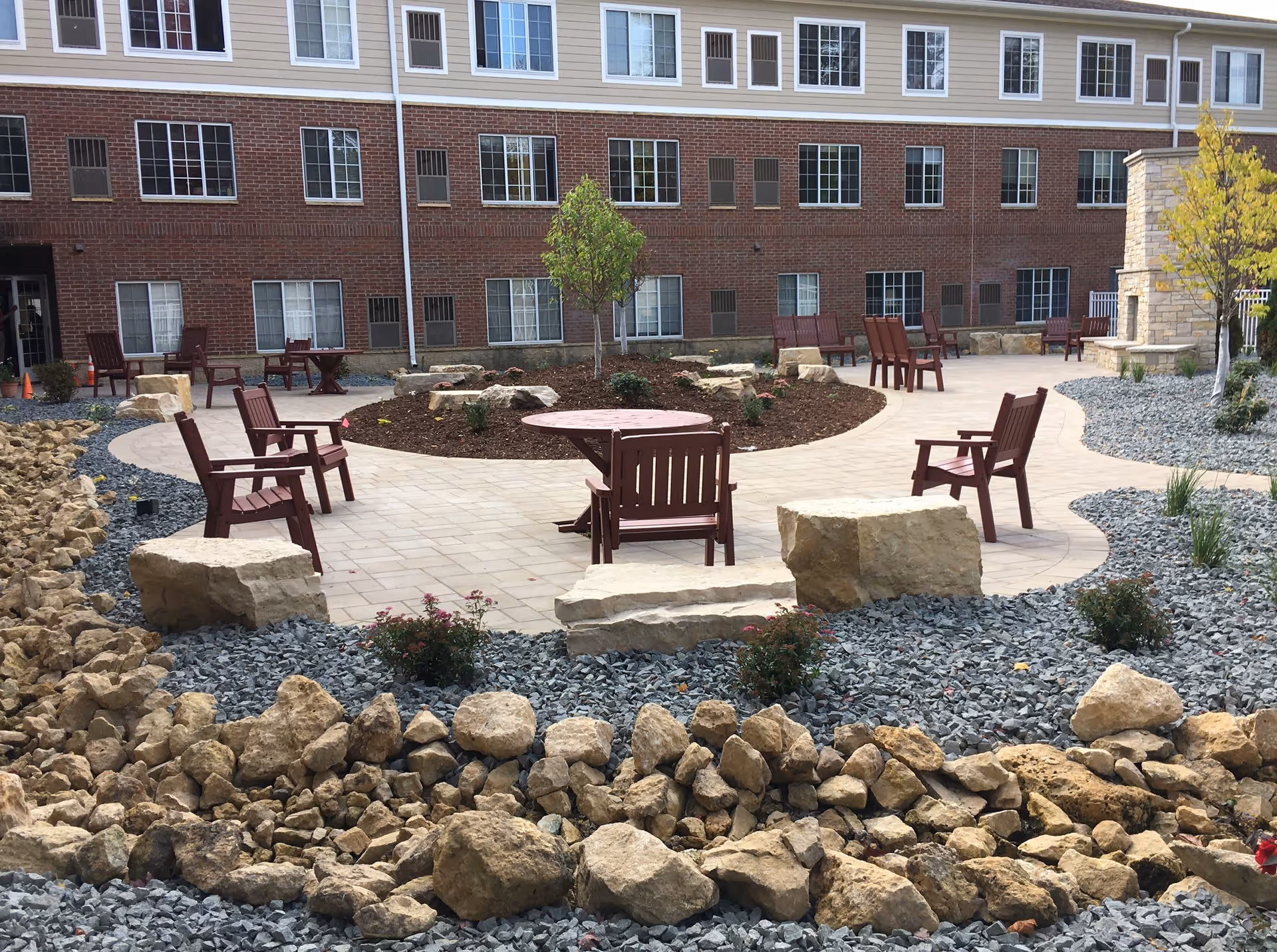 Outdoor courtyard area with a paved walkway, several wooden chairs and tables, large rocks, small trees, and a stone fireplace. The background shows a multi-story brick and beige building with many windows.
