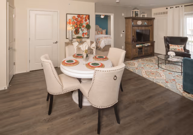 A cozy interior space featuring a round dining table set with four beige upholstered chairs. The table is set with plates, napkins, and a small floral centerpiece. In the background, there is a living area with a patterned rug, a wooden entertainment center with a TV, a dark leather armchair, and a glass coffee table. A bedroom with a bed and floral bedding is visible through an open doorway. The room has wood flooring and neutral-colored walls with a colorful floral painting hanging near the dining area.