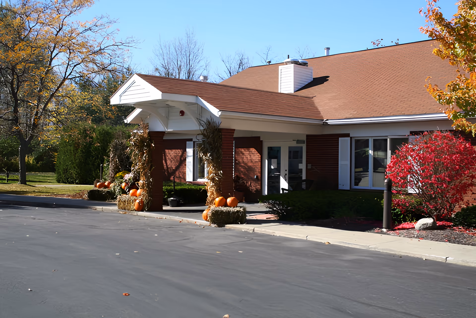 Exterior view of the entrance to Millview of Latham facility decorated with pumpkins and hay bales, surrounded by trees with autumn foliage under a clear blue sky.