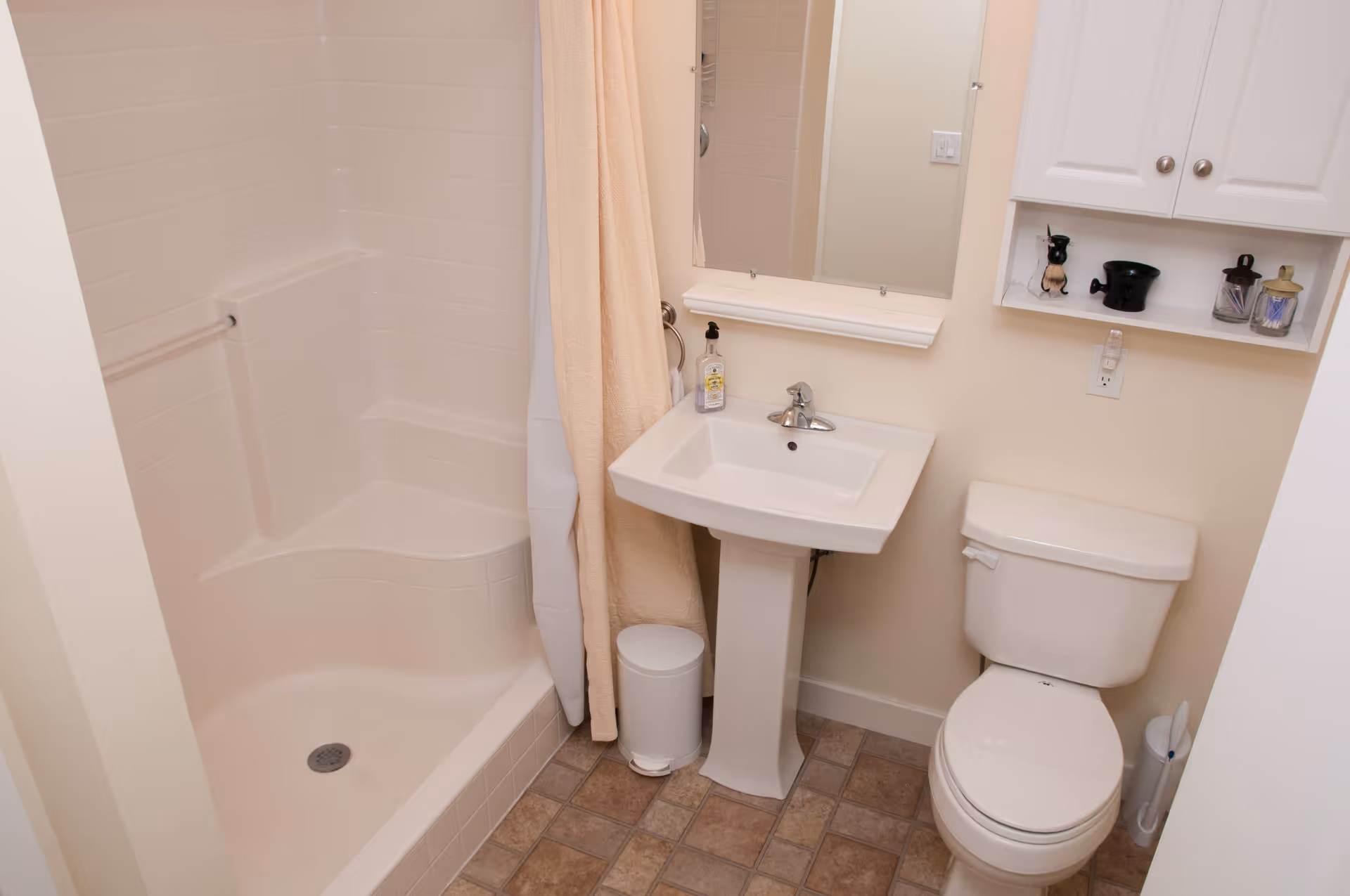 Compact bathroom featuring a shower with a curtain, a pedestal sink beneath a mirror, and a toilet under a wall cabinet.