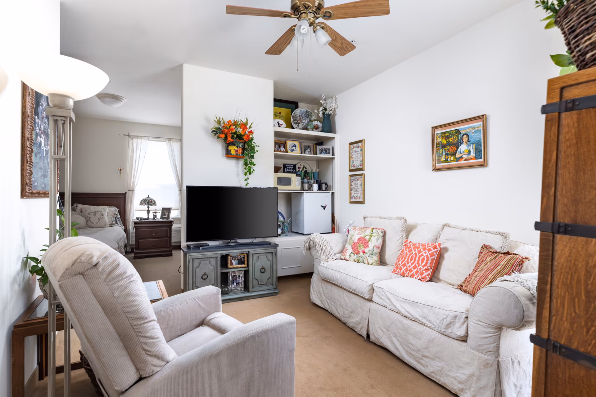 A cozy living room area with a beige recliner chair and a white sofa adorned with colorful cushions. A flat-screen TV sits on a vintage-style cabinet against a white wall with a floral wall decoration above it. Built-in shelves next to the TV hold a microwave, mini fridge, and various decorative items. A ceiling fan with wooden blades is mounted on the ceiling. In the background, a bedroom with a wooden bedframe and nightstand is visible through an open doorway.