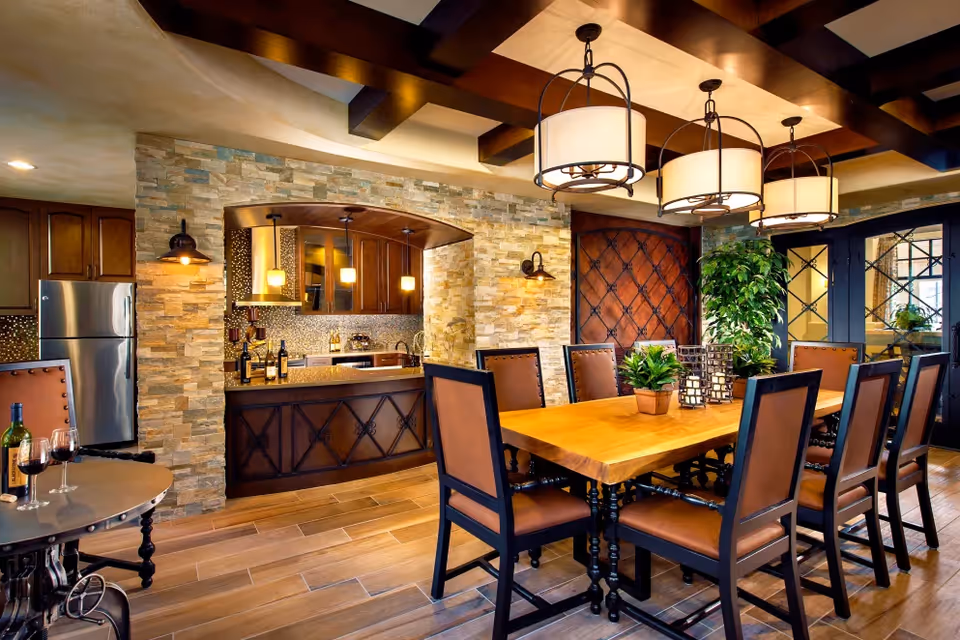 A warm and inviting dining room with a large wooden table surrounded by eight cushioned chairs. Above the table, three pendant lights hang from a ceiling with exposed wooden beams. In the background, there is a kitchen area with stone walls, wooden cabinets, a stainless steel refrigerator, and a countertop with wine bottles and glasses. There are also decorative plants on the table and near the wall.