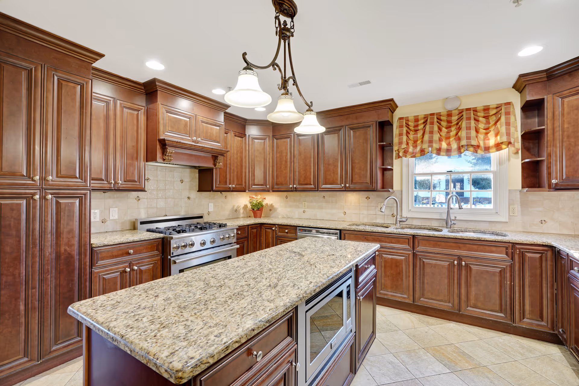 A spacious kitchen with dark wooden cabinets, granite countertops, a central island with a built-in microwave, a stainless steel gas stove, and a window with a red and yellow checkered valance.