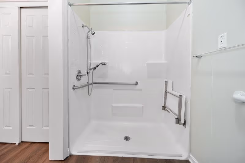 A clean, white walk-in shower with a handheld showerhead, grab bars on the walls, and a fold-down seat. The shower has a white tiled interior and a curtain rod above. The floor outside the shower is wood, and there are white double doors to the left.