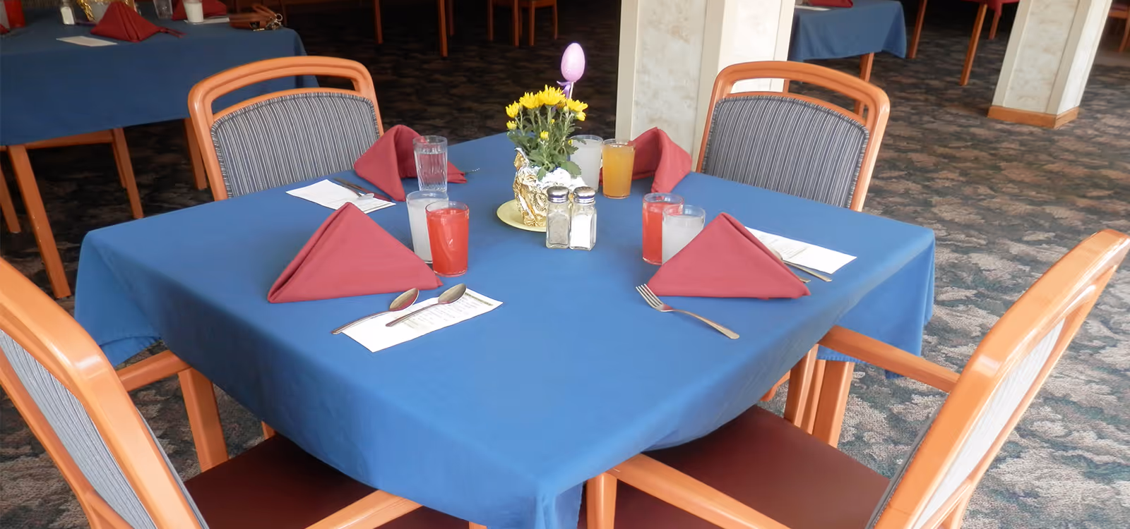 A dining table set for four with a blue tablecloth, red folded napkins, glasses of juice and water, silverware, menus, and a small floral centerpiece with yellow flowers and a decorative egg in a senior living facility.